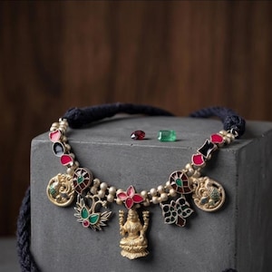 Può includere: Collana dorata con cordino blu scuro. La collana presenta vari ciondoli con dettagli smaltati rossi, verdi e neri. Una piccola statua dorata di una divinità è al centro della collana. Due pietre preziose, un rubino rosso e uno smeraldo verde, sono accanto alla collana.