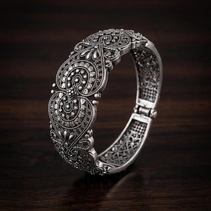 Può includere: Un braccialetto a bracciale incernierato color argento con intricati disegni floreali e a volute. Il braccialetto presenta un motivo dettagliato di forme vorticose e piccoli accenti circolari. La chiusura è visibile sul lato destro.