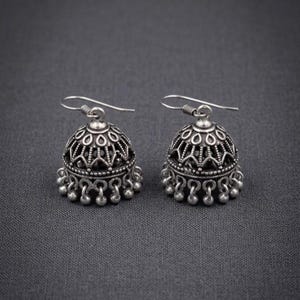 Può includere: Un paio di orecchini jhumka color argento. Ogni orecchino presenta un design a forma di campana con intricati motivi traforati e piccole perline pendenti lungo il bordo inferiore. Chiusura a gancio.