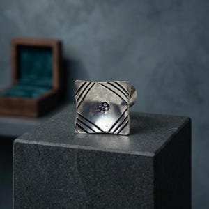 Könnte beinhalten: Ein quadratischer Silberring mit geometrischem Muster. Der Ring hat ein zentrales kreisförmiges Design, umgeben von diagonalen Linien. Er wird auf einer dunkelgrauen Oberfläche präsentiert, mit einer Holzschatulle im Hintergrund.