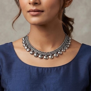 May include: A silver-toned choker necklace featuring a central braided chain design adorned with a series of small, round, polished metallic discs hanging along the bottom edge, complemented by a row of tiny silver beads along the top border.