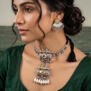 May include: Silver-toned necklace and matching earrings set. The necklace features a central pendant with a red stone and a gold-colored accent, with a strand of white pearls. The earrings have a fan-shaped design with gold and silver details.