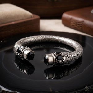 Könnte beinhalten: Ein silberfarbenes Armreif-Armband mit aufwendigen floralen Mustern und schwarzen Edelstein-Akzenten. Das offene Design hat einen Klappverschluss mit dekorativen Endkappen. Das Armband wird auf einer reflektierenden schwarzen Oberfläche präsentiert.