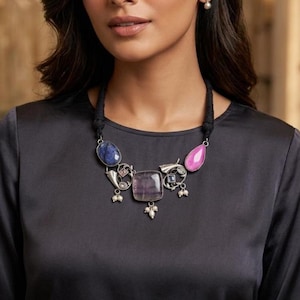 Könnte beinhalten: Eine auffällige Halskette mit schwarzem Kordel und silberfarbenen Metallelementen. Sie zeigt einen blauen Ovalstein, einen quadratischen lila Stein und einen rosa Tropfen-Edelstein. Kleine weiße Perlen akzentuieren das Design.
