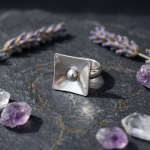 Könnte beinhalten: Ein silberner Ring mit quadratischer Fassung und erhabener Kugel in der Mitte. Der Ring liegt auf einer dunklen, strukturierten Oberfläche, im Hintergrund Lavendelzweige und Amethystkristalle. Das Design des Rings ist minimalistisch.