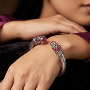 Könnte beinhalten: Silbernes Armreif-Armband mit aufwendigen Details und drei ovalen, roten Edelsteinen an jedem Ende. Das offene Armband wird am Handgelenk getragen und zeigt das komplizierte Design und die leuchtende Farbe der Steine.