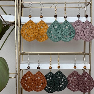 May include: A display of handmade crochet earrings in various colors, including mustard yellow, teal, and mauve. Each pair features a teardrop shape with a detailed floral pattern and wooden bead accents. The earrings hang from silver or gold-toned hooks.
