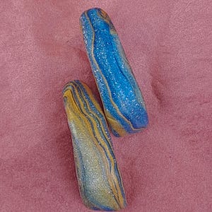 Peut inclure: Deux pierres décoratives oblongues à effet marbré. Les pierres sont principalement bleues et jaunes, avec une finition scintillante et mouchetée. Le texte "Longueur: 3,5 cm" est visible sur l'image. Les pierres mesurent environ 3,5 cm de long.