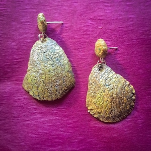 Puede incluir: Un par de pendientes hechos a mano con un diseño texturizado y orgánico. Los pendientes presentan una paleta de colores dorados y verdes con un patrón similar al de un reptil. Los pendientes se exhiben sobre un fondo púrpura.