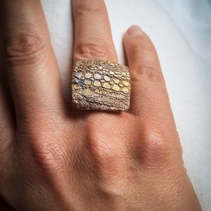 Può includere: Anello quadrato strutturato con un motivo a mosaico. L'anello presenta tonalità oro, argento e bronzo, creando un design unico. L'anello è indossato su un dito. Il design dell'anello ricorda una texture di corteccia.