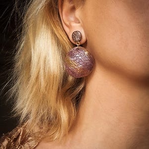 May include: A close-up of an ear adorned with statement earrings. The earrings have a textured, purple and gold spherical design, with a small, dark brown disc stud. The lower part of the earring is a larger, textured sphere.
