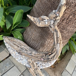 May include: A whale-shaped wall art piece constructed from weathered driftwood and small white seashells. The sculpture features a curved tail and a rounded body, with the driftwood forming the structure and the shells covering the lower section.