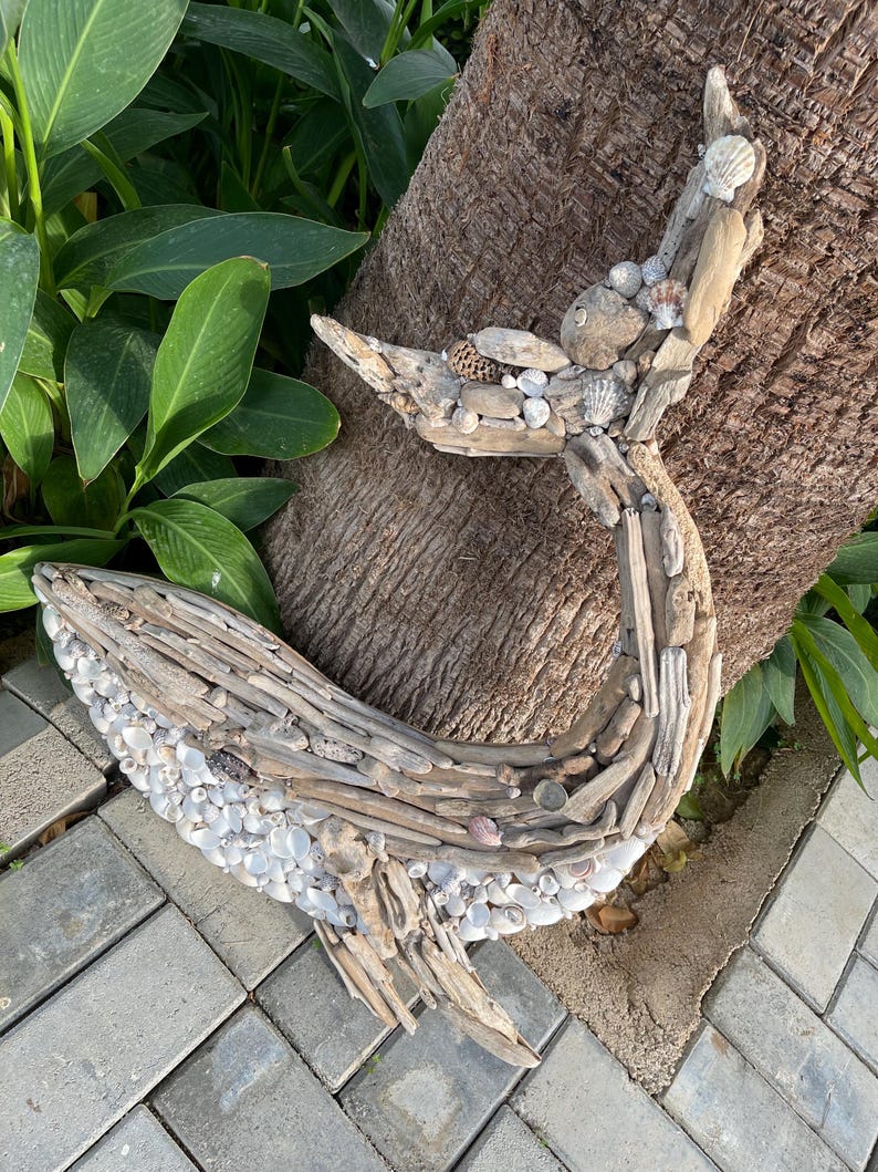 May include: A whale-shaped wall art piece constructed from weathered driftwood and small white seashells. The sculpture features a curved tail and a rounded body, with the driftwood forming the structure and the shells covering the lower section.