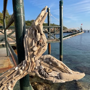 May include: A whale tail sculpture crafted from light-colored driftwood pieces and various small seashells. The textured artwork is assembled with organic materials, featuring a layered composition of weathered wood fragments and white sea shells.