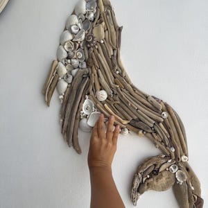 Op de afbeelding: Een handgemaakt kunstwerk voor aan de muur in de vorm van een walvis. Het kunstwerk is gemaakt van drijfhout en schelpen, met een mix van lichtbruine en witte tinten. De walvis is samengesteld uit kleine stukjes drijfhout.