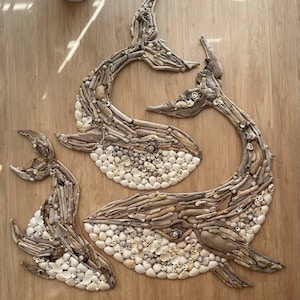 May include: Three whale-shaped wall art pieces constructed from light brown driftwood and various white and tan seashells. The arrangement features two larger whales positioned in an overlapping manner and one smaller whale placed to the left side.