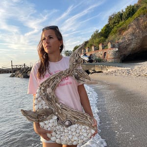 May include: A decorative wall art piece shaped like a whale, constructed from light-colored driftwood and a dense mosaic of small white seashells. The sculpture features a curved tail and a rounded body, with natural wood textures and varied shell sizes.