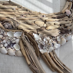 May include: A wall art piece shaped like a whale, constructed from various pieces of light brown driftwood and an assortment of white, cream, and grey seashells. The eye is formed by a dark, textured spiral shell, and the fins are made of wood.