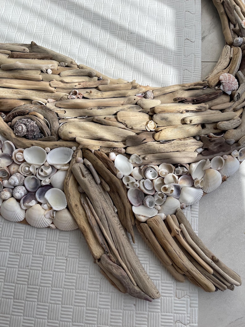 May include: A wall art piece shaped like a whale, constructed from various pieces of light brown driftwood and an assortment of white, cream, and grey seashells. The eye is formed by a dark, textured spiral shell, and the fins are made of wood.