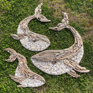 May include: Three whale-shaped wall art pieces crafted from light-colored driftwood and small white seashells. Each whale figure features a curved tail, a distinct eye made from a dark shell, and a textured body composed of layered wood and shells.