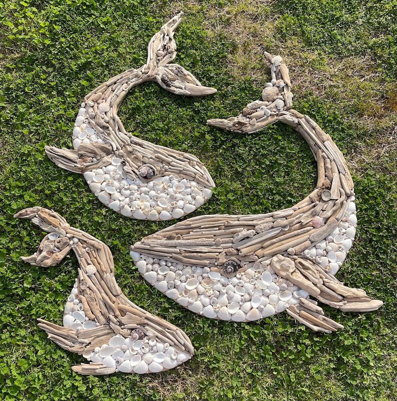 May include: Three whale-shaped wall art pieces crafted from light-colored driftwood and small white seashells. Each whale figure features a curved tail, a distinct eye made from a dark shell, and a textured body composed of layered wood and shells.
