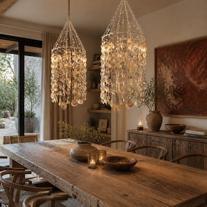 May include: Two shell chandeliers hang above a rustic wooden dining table. The chandeliers have a conical shape, with shells cascading down. The table is surrounded by wooden chairs. A wooden cabinet and artwork are visible in the background.