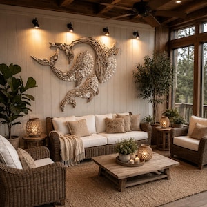 May include: A large wall art piece made of driftwood and white seashells shaped like two whales, mounted on a white vertical paneled wall above a brown wicker sofa with white cushions, throw pillows, and a cream-colored knit throw blanket.