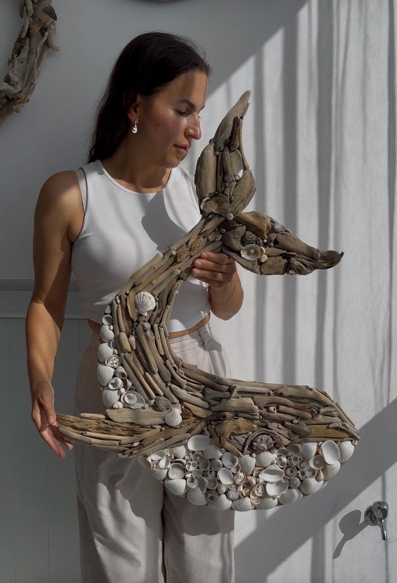 May include: A wall art sculpture shaped like a whale tail, constructed from various pieces of weathered driftwood and an assortment of small white seashells. The natural wood tones and white shells create a textured, organic surface on the whale tail.