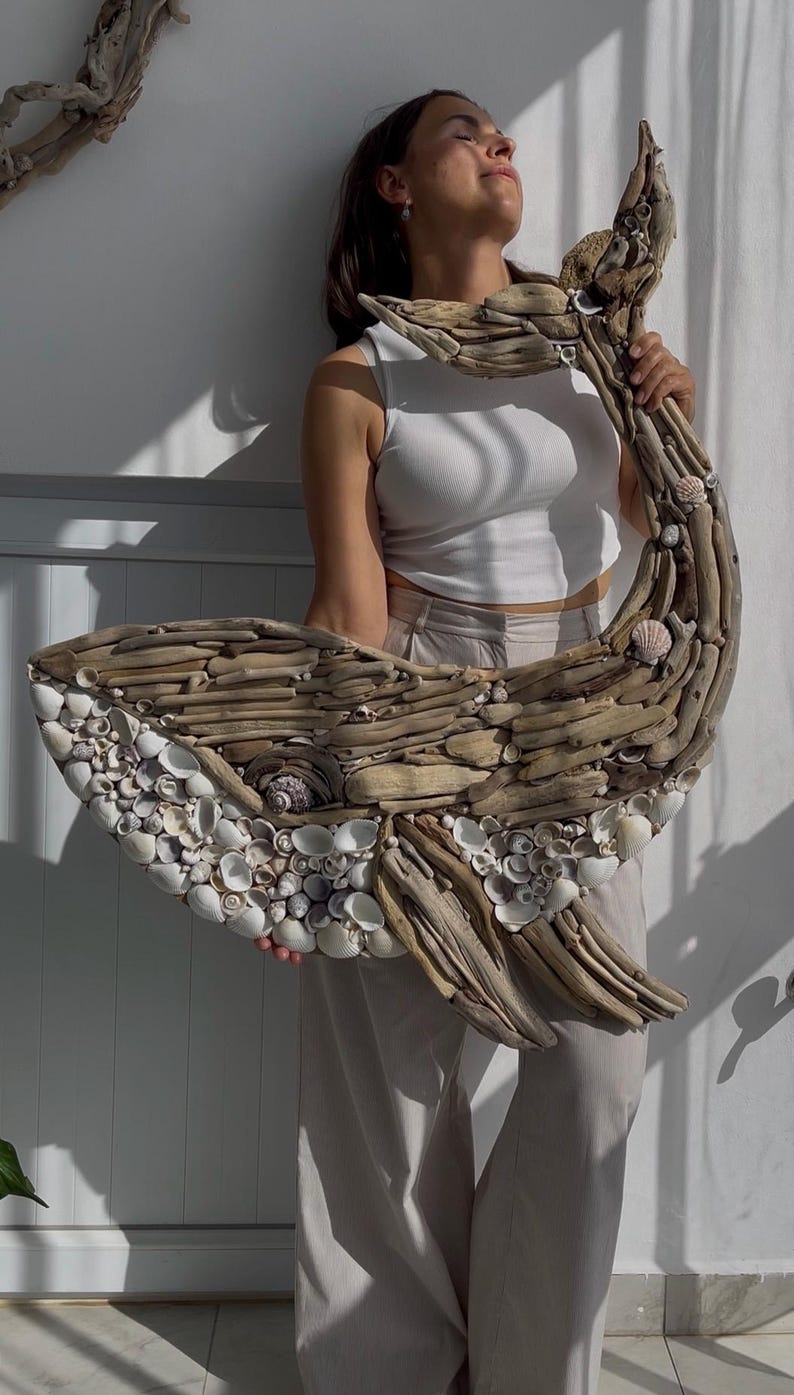 May include: A large wall sculpture shaped like a whale, constructed from various pieces of weathered driftwood and small white seashells. The driftwood forms the body and tail, while the white shells create the underbelly and eye detail.