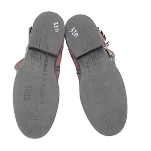 May include: Pair of brown leather ankle boots with dark gray soles. The soles have embossed patterns and text. The number "320" is written in white on the heel of each boot. The boots have buckles and fringe.