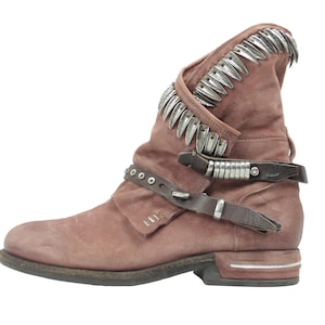 May include: A dusty rose-colored ankle boot with a distressed finish. The boot features a decorative silver metal trim along the top edge and straps with silver accents. The boot has a low heel and a rounded toe.