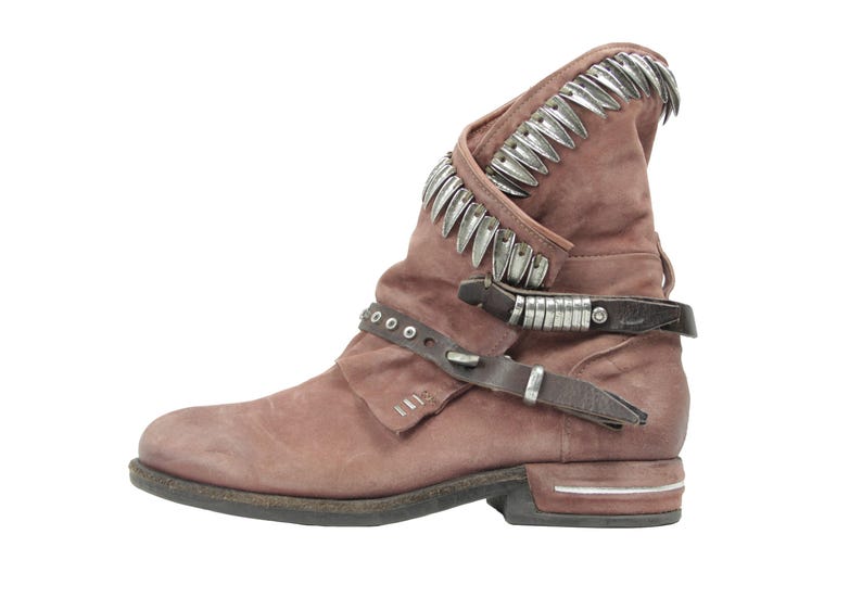 May include: A dusty rose-colored ankle boot with a distressed finish. The boot features a decorative silver metal trim along the top edge and straps with silver accents. The boot has a low heel and a rounded toe.