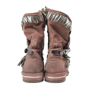 May include: Pair of brown suede boots with silver-tone metal accents along the top and straps. The boots have buckles and fringe details, with a zipper on the side. The soles have a silver stripe.