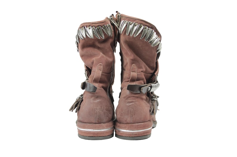 May include: Pair of brown suede boots with silver-tone metal accents along the top and straps. The boots have buckles and fringe details, with a zipper on the side. The soles have a silver stripe.