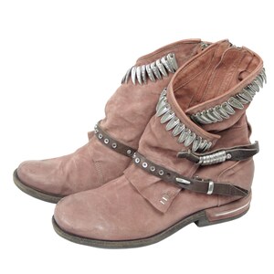 May include: A pair of dusty rose-colored ankle boots with a distressed finish. The boots feature silver-toned decorative trim around the top and a brown leather strap with silver studs and a buckle. The boots have a low heel and a side zipper.