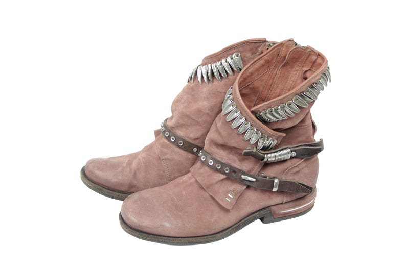 May include: A pair of dusty rose-colored ankle boots with a distressed finish. The boots feature silver-toned decorative trim around the top and a brown leather strap with silver studs and a buckle. The boots have a low heel and a side zipper.