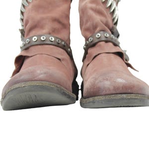 May include: A pair of dusty rose-colored boots with a distressed appearance. The boots feature decorative silver accents at the top and studded leather straps around the ankles. The soles are dark gray.