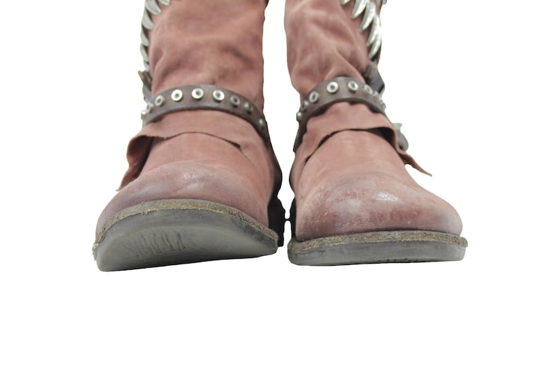 May include: A pair of dusty rose-colored boots with a distressed appearance. The boots feature decorative silver accents at the top and studded leather straps around the ankles. The soles are dark gray.