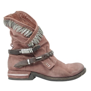 May include: Dusty rose-colored ankle boot with silver-toned metal accents. The boot features a folded-over top, two brown leather straps with buckles, and a low heel. The design includes decorative metal studs.