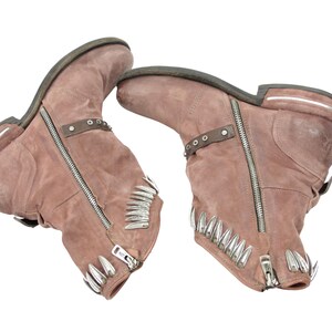 May include: A pair of dusty rose-colored ankle boots with silver zippers and decorative metal accents. The boots have a rugged, worn appearance, with a low heel and a strap detail around the ankle. The toe of each boot is adorned with silver spikes.