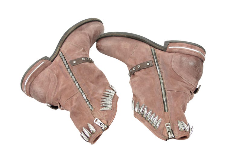 May include: A pair of dusty rose-colored ankle boots with silver zippers and decorative metal accents. The boots have a rugged, worn appearance, with a low heel and a strap detail around the ankle. The toe of each boot is adorned with silver spikes.