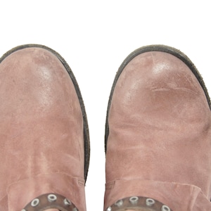 May include: Overhead shot of two dusty rose-colored leather boots. The boots have a distressed appearance with a dark brown sole and a decorative metal accent near the ankle. The boots are set against a white background.