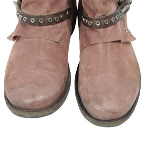 May include: Pair of dusty rose-colored ankle boots. The boots have a distressed leather appearance with a dark brown strap around the ankle, adorned with silver-colored rivets. The soles are dark with a worn look.