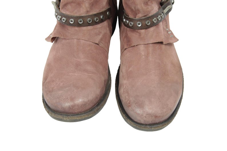 May include: Pair of dusty rose-colored ankle boots. The boots have a distressed leather appearance with a dark brown strap around the ankle, adorned with silver-colored rivets. The soles are dark with a worn look.