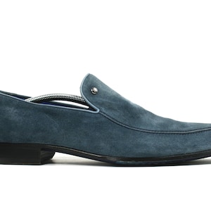 May include: A blue suede loafer with a low heel and a rounded toe. The shoe has a stitched design around the top and a small silver-colored button detail. A shoe tree is inside the shoe.