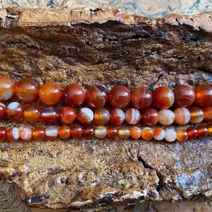 May include: Three strands of gemstone beads in shades of orange, red, and white. The beads are round and vary in size, with the largest beads at the top. The beads are displayed on a textured, brown surface.