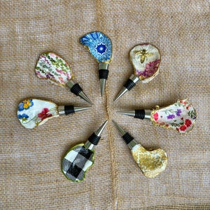 May include: A selection of decorative bottle stoppers with various designs. Each stopper showcases a different pattern, such as floral, geometric, and bee motifs. The stoppers have a silver metal base and a black rubber seal.