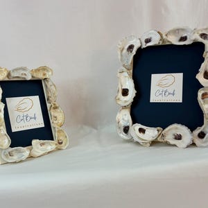 May include: Two white picture frames with navy blue inserts, decorated with oyster shells. The frames display the "Cat Bank Inspirations" logo. One frame is upright, the other angled, on a white surface.
