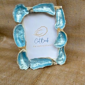 May include: A rectangular photo frame decorated with oyster shells. The shells are painted with a pearlescent light blue colour and are arranged around the frame. The frame has the text "Cut Bank Inspirations".
