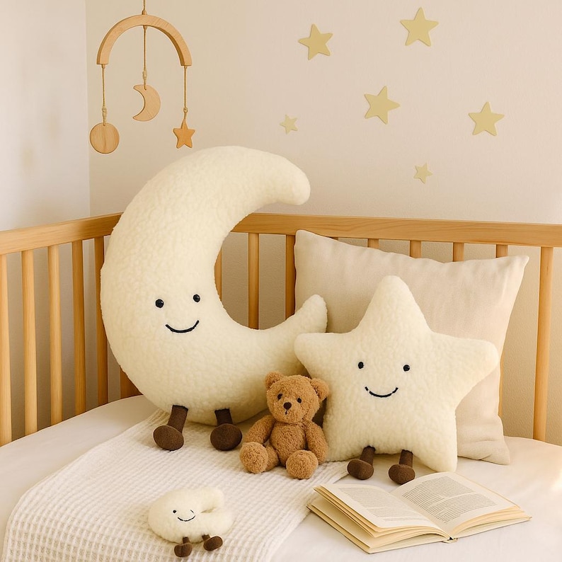 May include: A nursery scene with a cream-colored crescent moon and star-shaped plush pillows with smiling faces. A small teddy bear and a cloud-shaped pillow are also present. A wooden mobile hangs above the crib, and star-shaped wall decorations complete the scene.